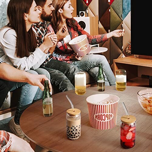 Group of friends watching TV with popcorn and drinks on table.