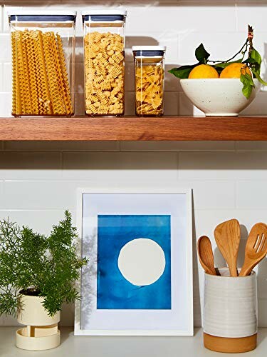 Kitchen counter with pasta jars, fruit bowl, artwork, plant, and utensils.